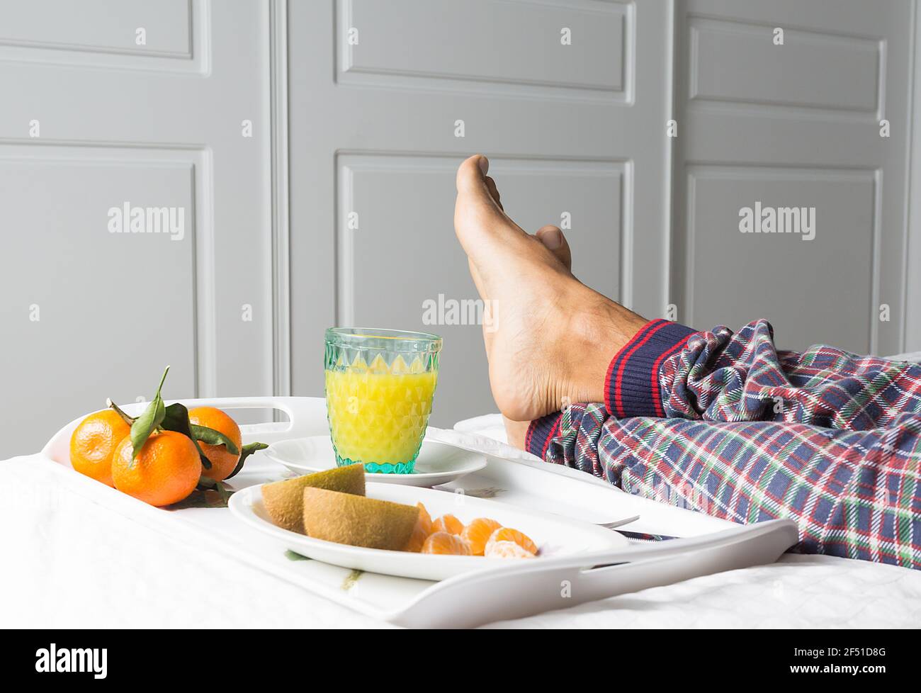 Imagen horizontal de las piernas de un hombre en pijama a cuadros parte  superior de una cama con una cubierta blanca junto a. una bandeja de  desayuno con fruta y zumo de