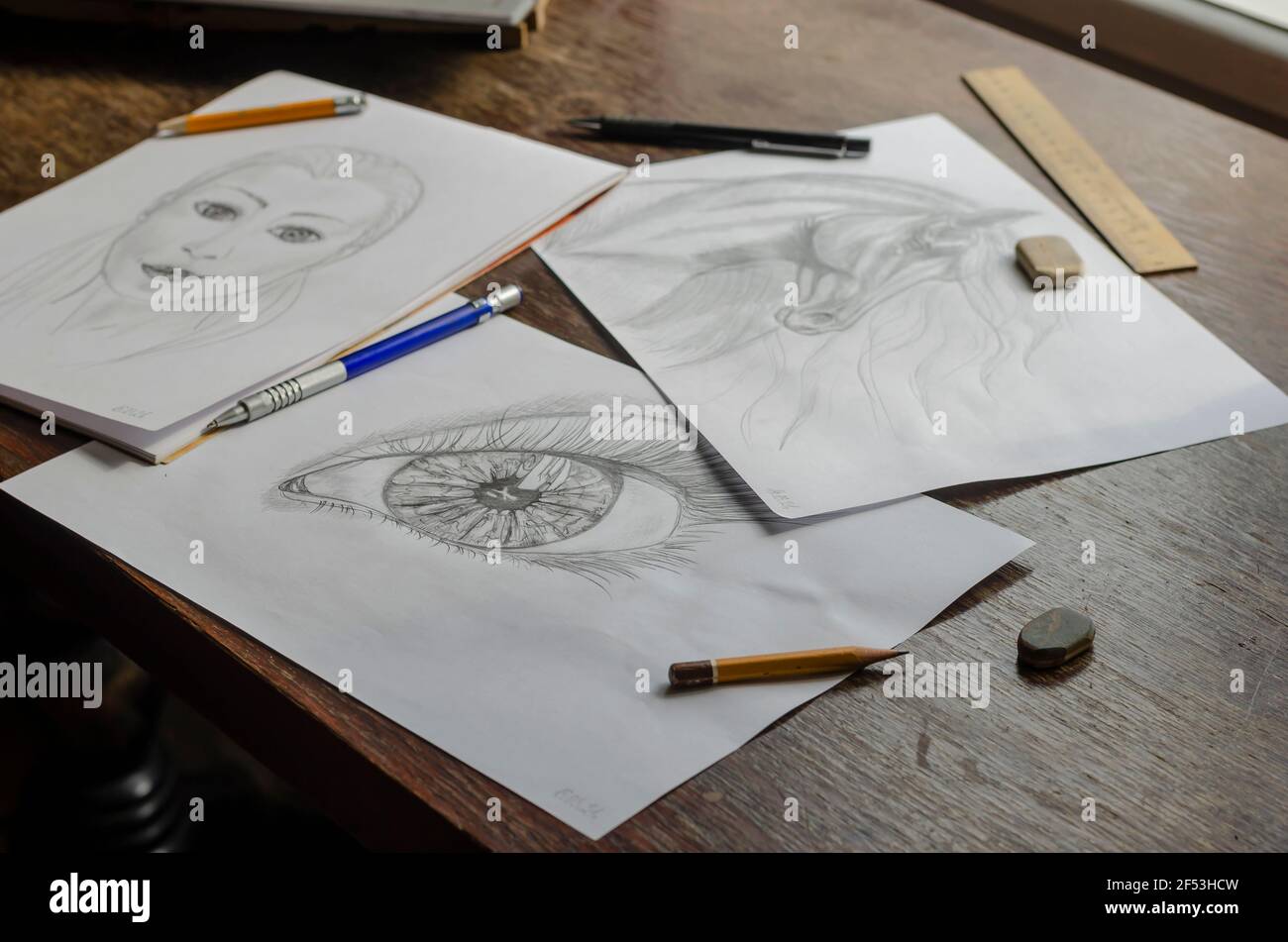 Los dibujos hechos con un lápiz se encuentran sobre una mesa de madera  marrón. Caballo, ojo humano y retrato conceptual femenino dibujado en papel  blanco. Lápices, borrador y regla Fotografía de stock -