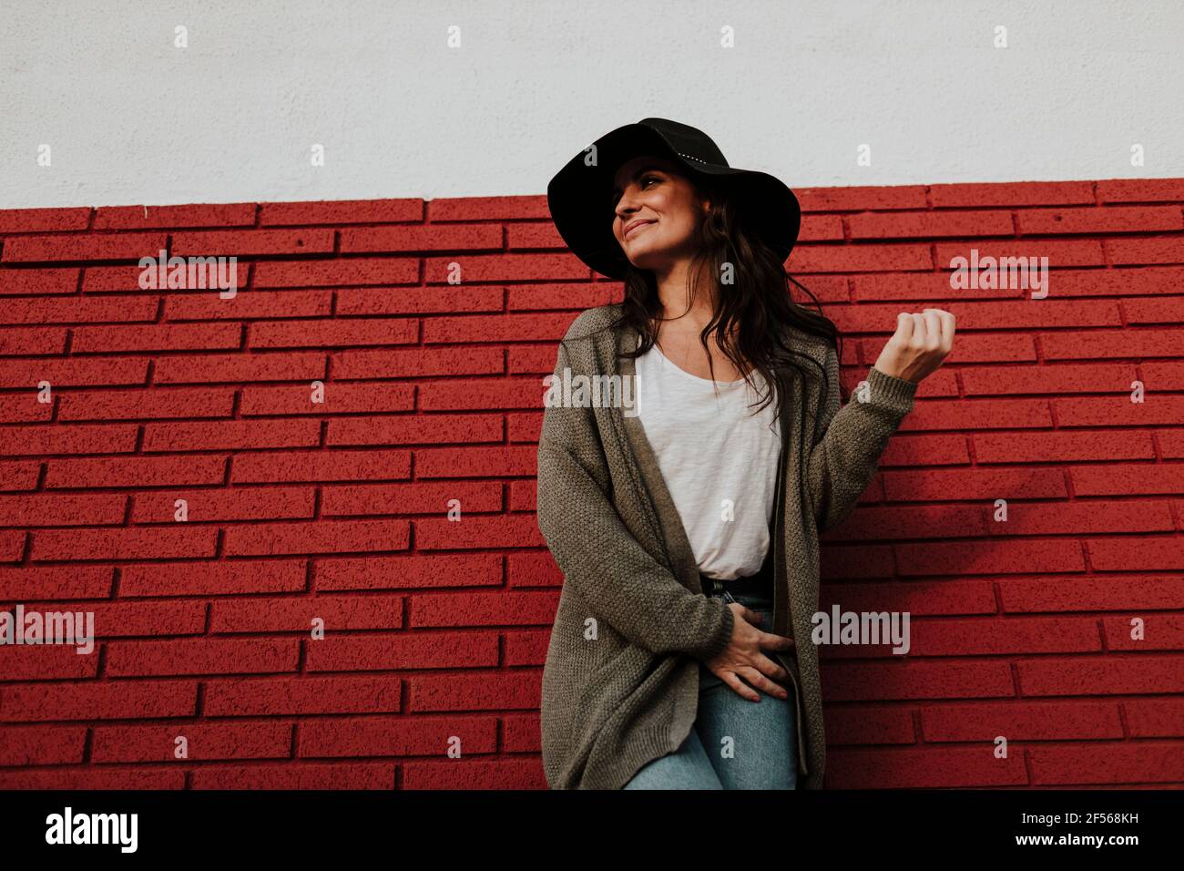 Mujer en el sombrero mirando lejos mientras que parada contra la pared roja del ladrillo Foto de stock