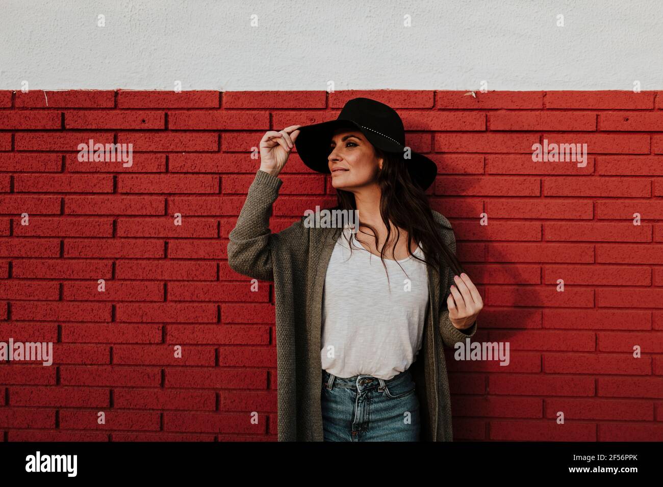 Mujer sosteniendo sombrero con los ojos cerrados de pie contra la pared de ladrillo rojo Foto de stock