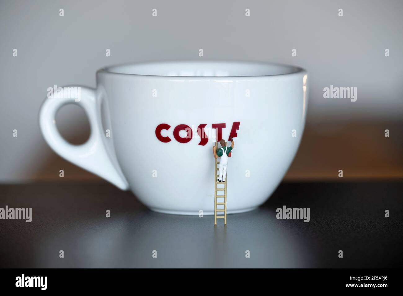 Una pequeña figura de pintor y decorador en miniatura a escala 00 se muestra usando una escalera para pintar el logotipo de la marca Costa Coffee en una taza de café costa. Foto de stock
