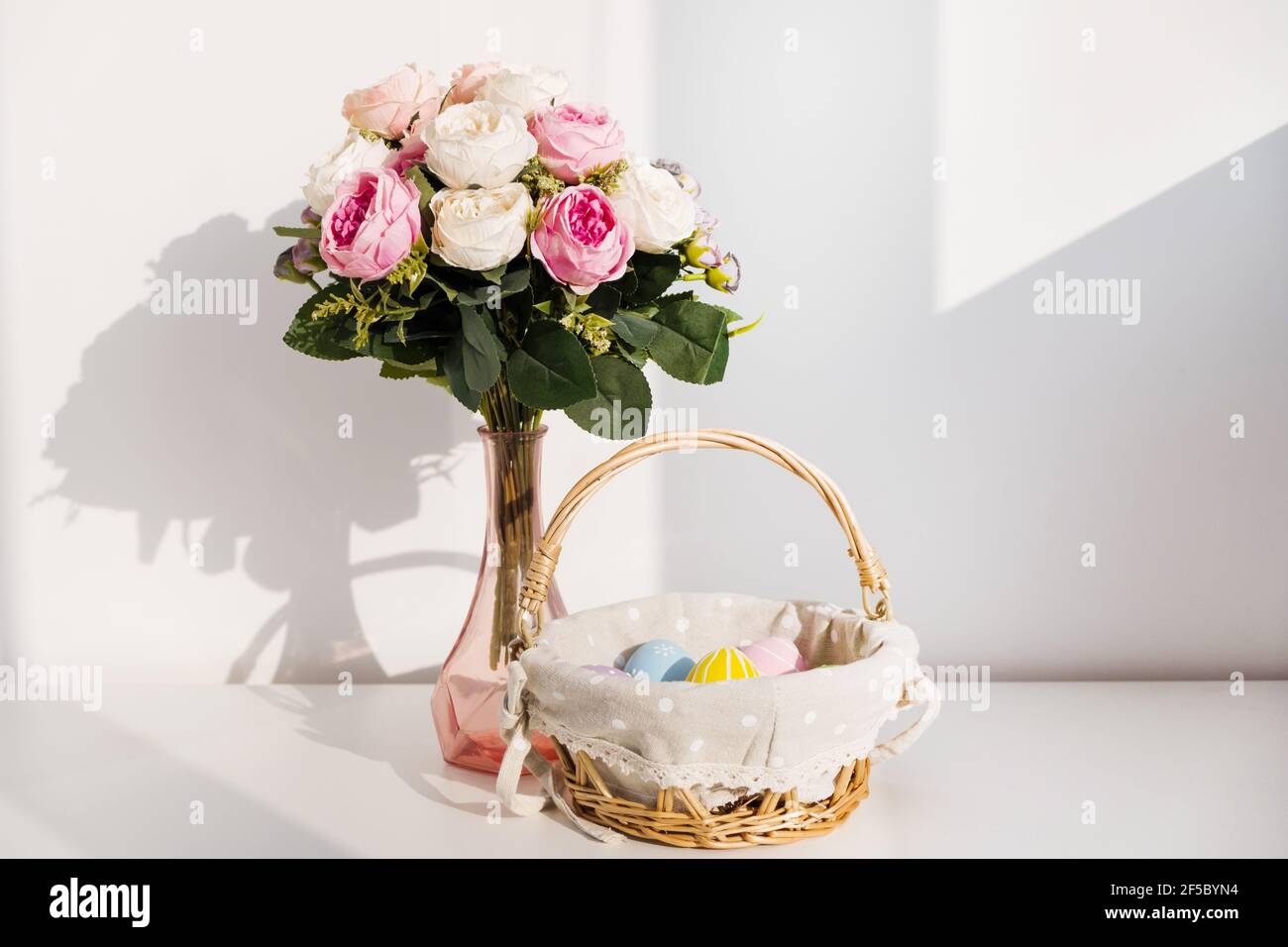 Huevos coloridos de Pascua en buket sobre mesa de madera blanca. Ramo de flores de rosas en el fondo. Felices Pascuas. Estado de ánimo de primavera. Foto de stock