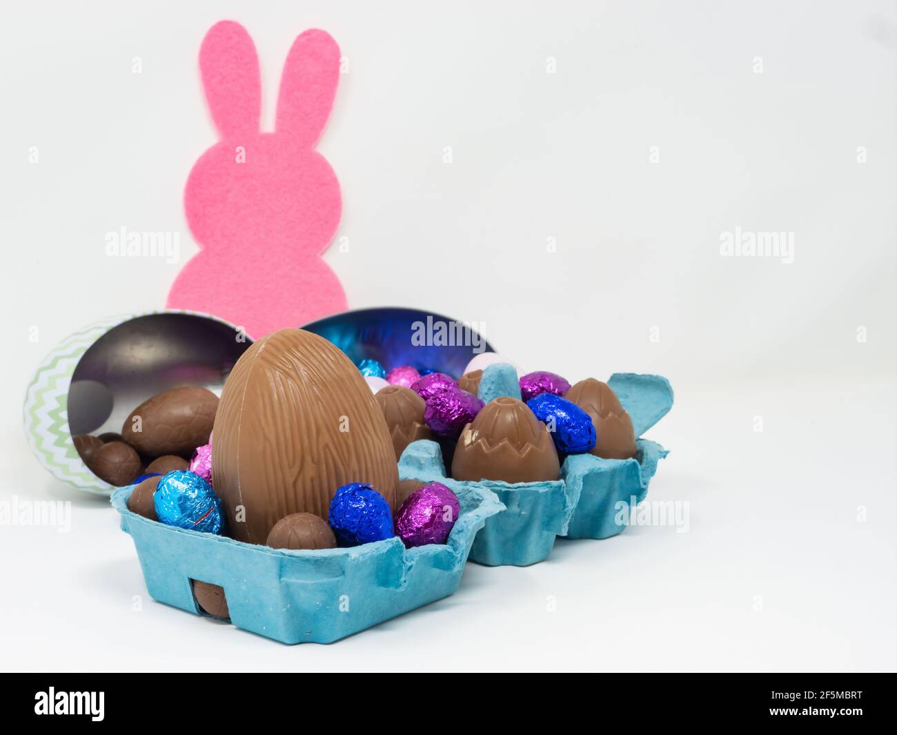 Un montón de huevos de Pascua de chocolate de diferentes tamaños en un cartón de huevo y en recipientes con forma de huevo sobre fondo blanco con una silueta de conejito rosa Foto de stock