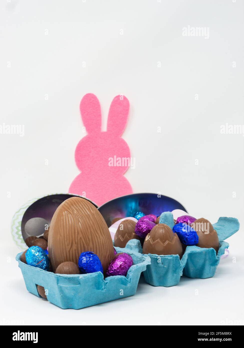 Un montón de huevos de Pascua de chocolate de diferentes tamaños en un cartón de huevo y en recipientes con forma de huevo sobre fondo blanco con una silueta de conejito rosa Foto de stock