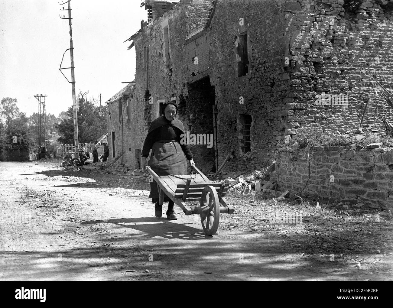 Francia rural Segunda Guerra Mundial 1944. La vieja dama empujando el carro de mano a través de la aldea francesa pasado la guerra dañó los edificios con soldados aliados en el fondo durante la liberación de Francia Foto de stock