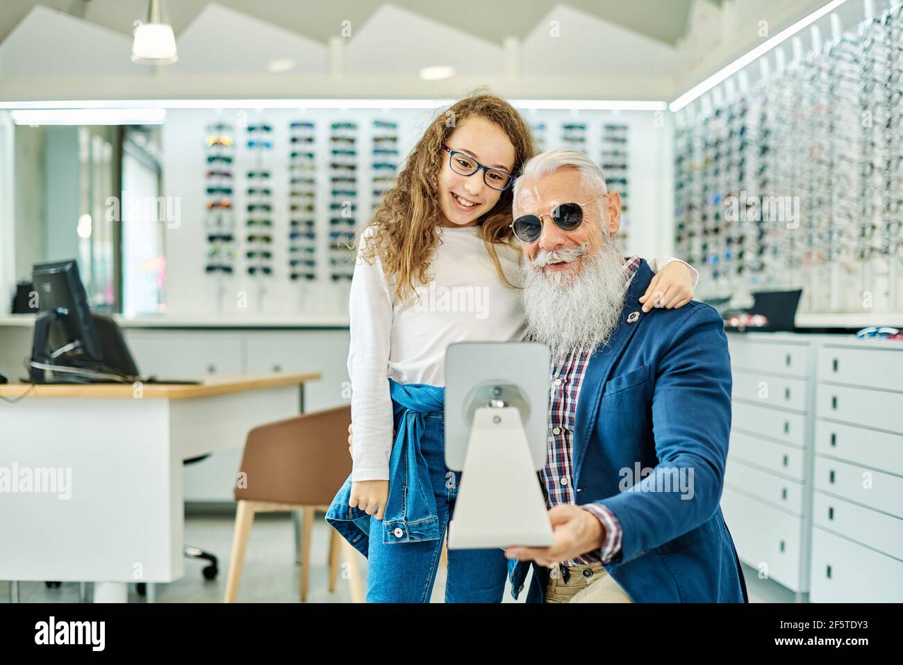 Alegre hombre mayor sosteniendo el espejo mientras la adolescente lo gafas en una tienda óptica moderna Fotografía de - Alamy