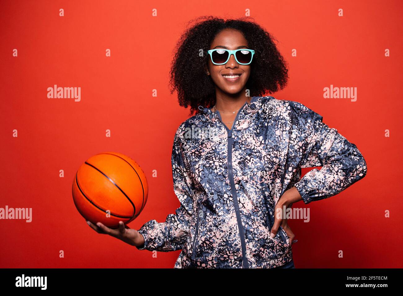 Mujer afroamericana de moda en estilo urbano y ropa fresca gafas de sol de pie con pelota baloncesto en las manos contra fondo rojo Fotografía de stock - Alamy