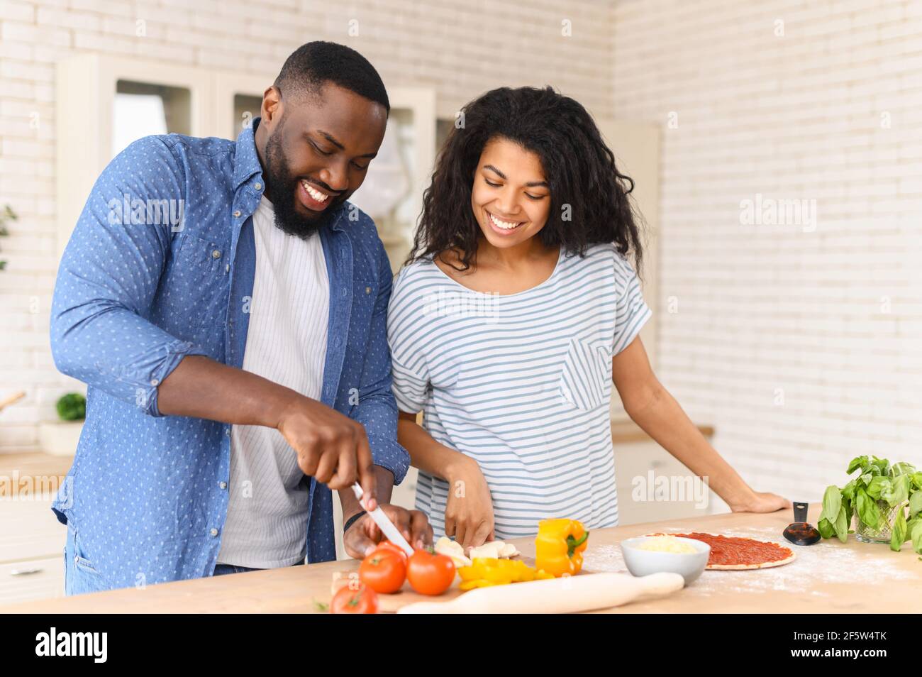 La joven familia feliz prepara su primer plato juntos en un nuevo apartamento, marido corta el queso para pizza, pararse en la mesa con muchas verduras y la masa preparada impregnada con pasta de tomate Foto de stock