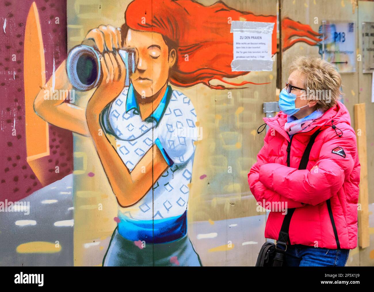 Münster, NRW, Alemania. 28th de marzo de 2021. Las personas que usan  máscaras pasan por una valla de construcción decorada con un dibujo de un  fotógrafo que golpea a los peatones en