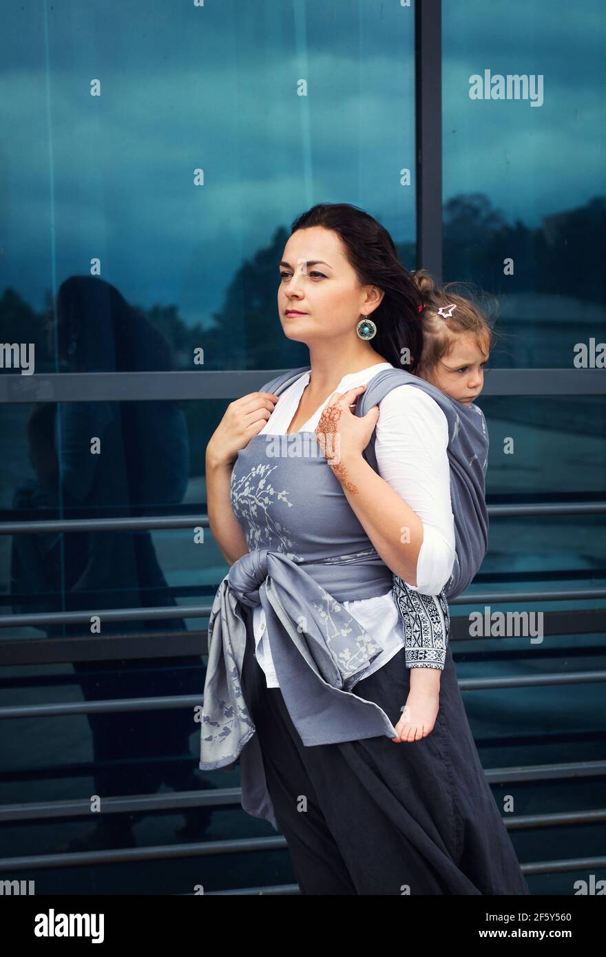 La madre y la niña se sientan en un cabestrillo gris permanezca cerca del cristal urbano Foto de stock