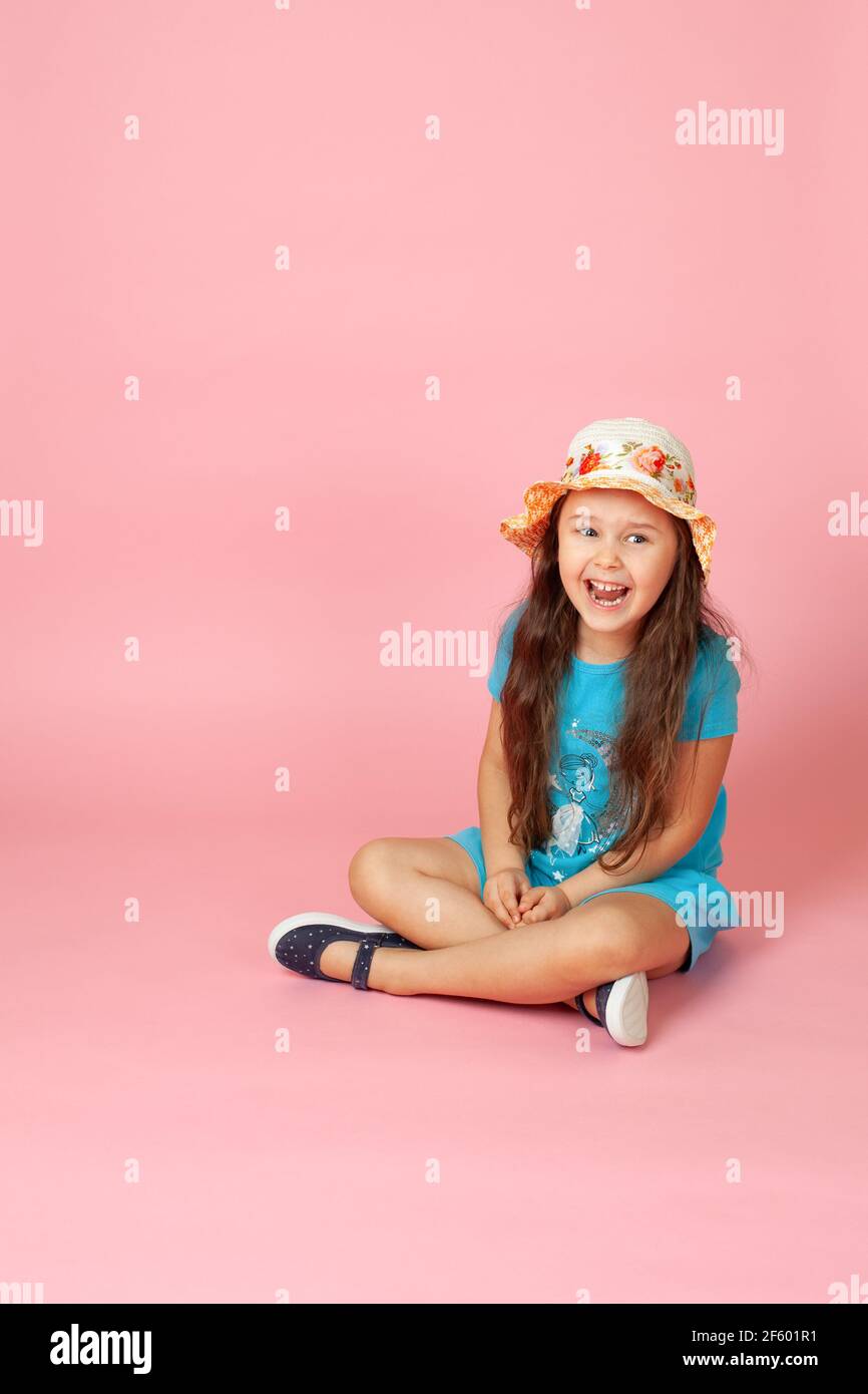 chica de boca abierta con pelo largo y ondulado en un sombrero de paja y vestido azul sentado con patas cruzadas en el suelo, aislado sobre un fondo rosa Foto de stock