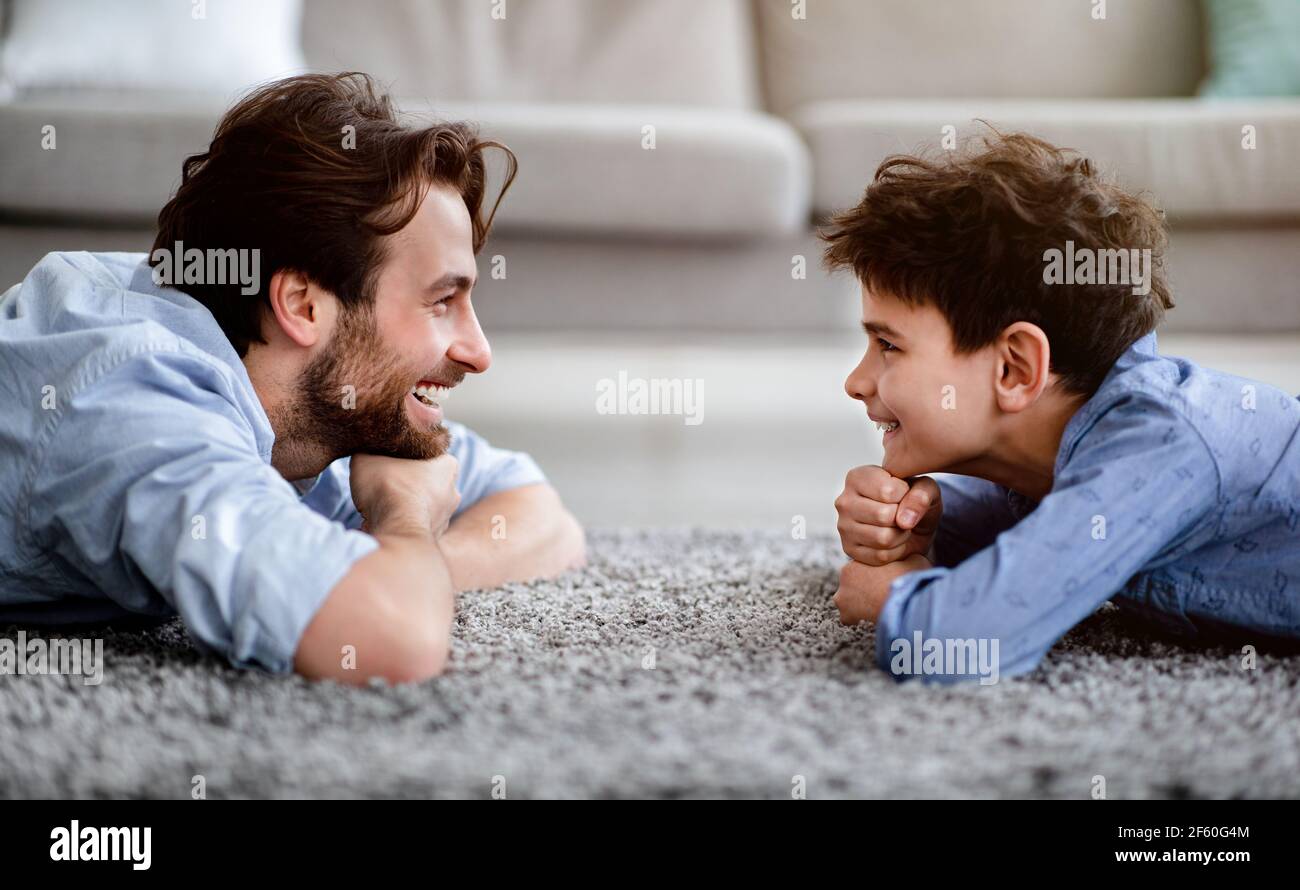 Dos generaciones. Perfil retrato de padre feliz y su hijo acostado en la alfombra en casa y mirando el uno al otro, vista lateral Foto de stock