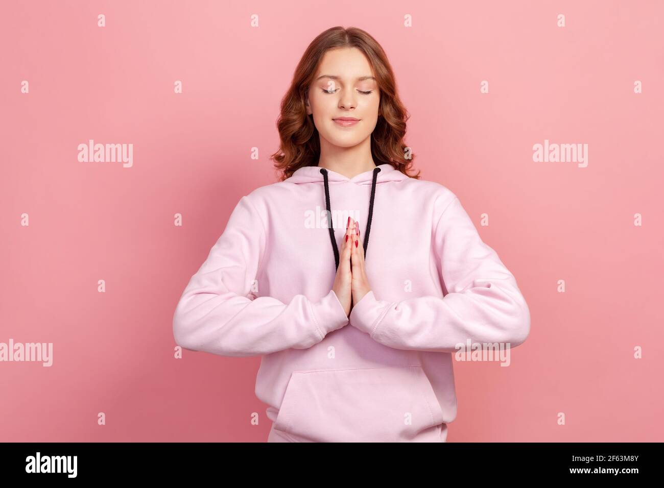 Retrato de niña tranquila en hoodie concentrando su mente, manteniendo las manos gesto namaste, meditando, yoga ejercicio respiración técnica reducir el estrés. Foto de stock