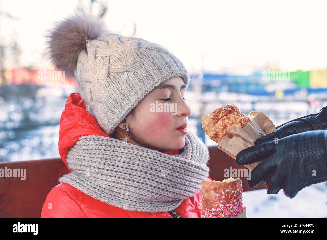 Joven hermosa chica caucásica vestida con una chaqueta brillante comiendo un pastel colorido. Actividad al aire libre en invierno, infancia feliz. Foto de stock