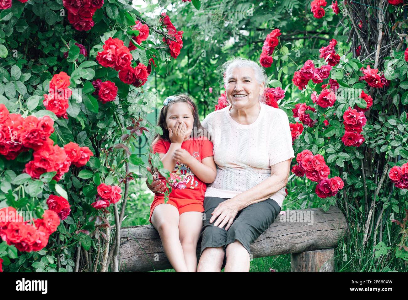 una anciana de pelo blanco con una sonrisa blanca como la nieve y una una niña de cinco años se divierte sentada en un banco el patio con rosales Foto de stock
