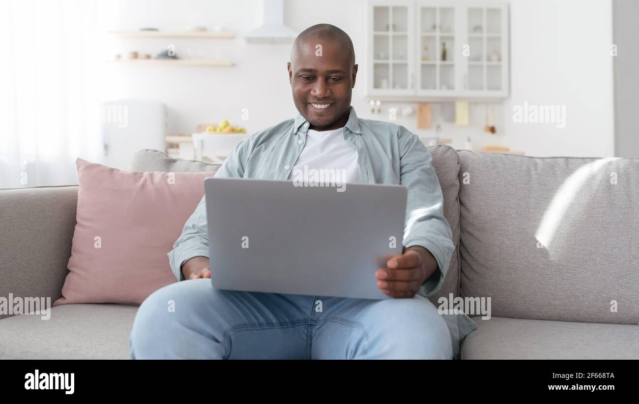 Feliz hombre negro de mediana edad utilizando el portátil mientras descansa en el sofá en la sala de estar, panorama Foto de stock
