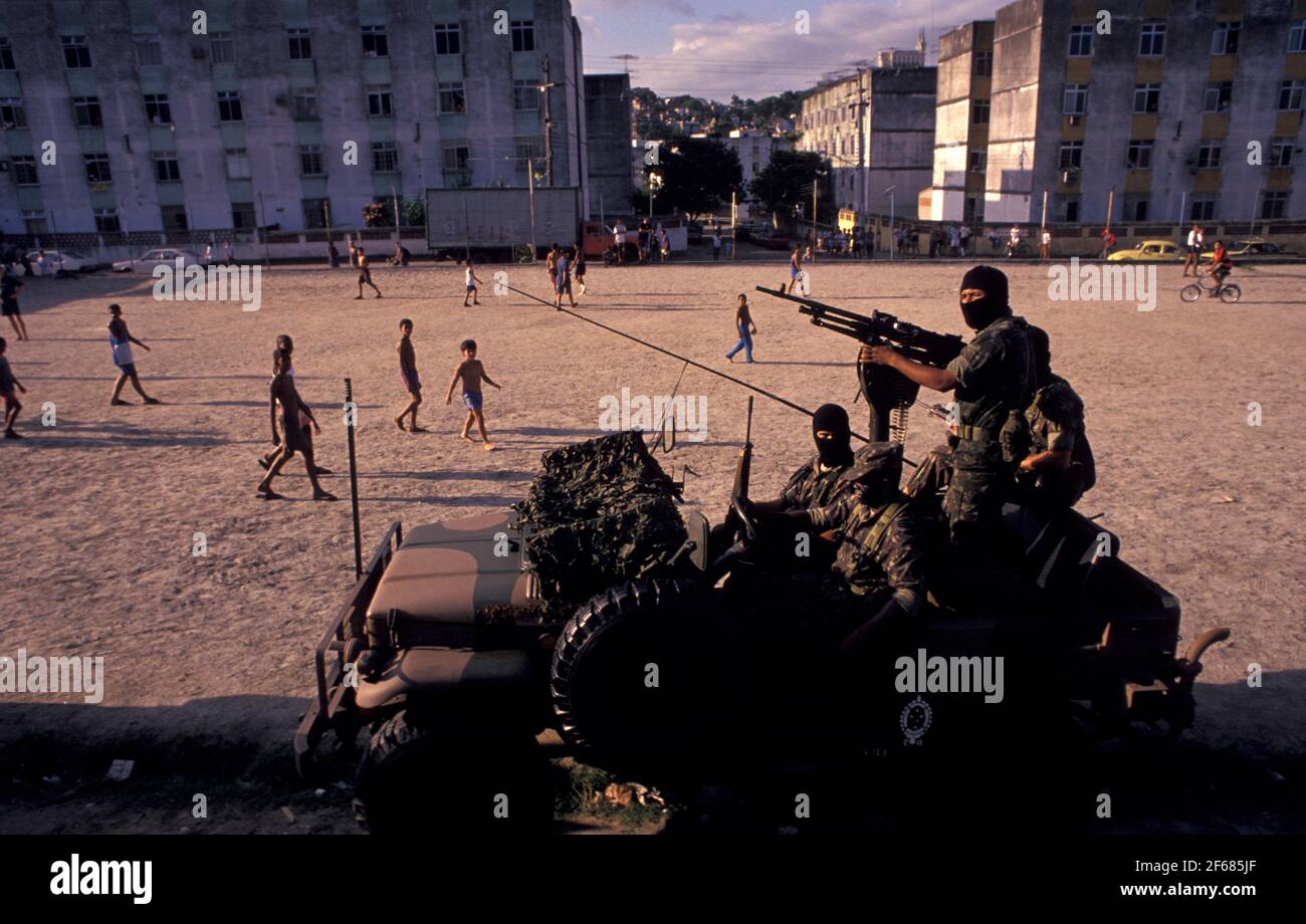Los jóvenes juegan al fútbol en un área empobrecida de Río de Janeiro mientras que una persona militar busca traficantes de drogas. Ocupación por el ejército de las zonas desfavorecidas como parte de la política gubernamental de seguridad pública en preparación de la Copa Mundial de la FIFA 2014 en Brasil. Foto de stock