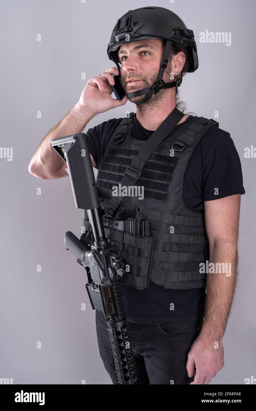 Hombre equipado para airsoft con casco y chaleco protector está confirmando la estrategia en el teléfono mientras que de pie con su arma mostrada. Negociación Foto de stock