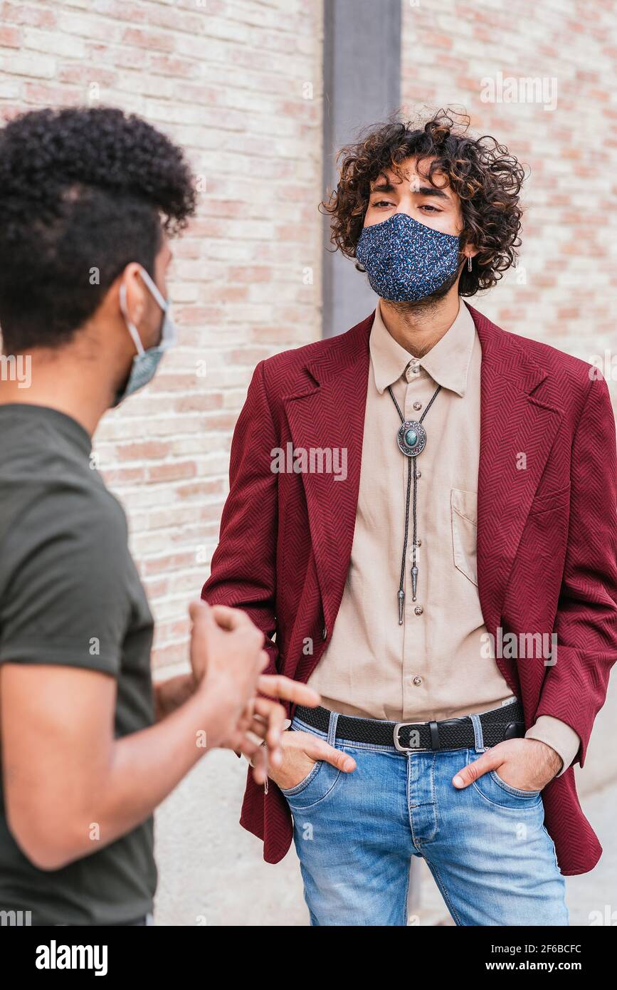 retrato de un joven caucásico vestiéndose ropa de moda y máscara azul. Está hablando con un colega latino en la calle cerca de una pared de ladrillo Foto de stock