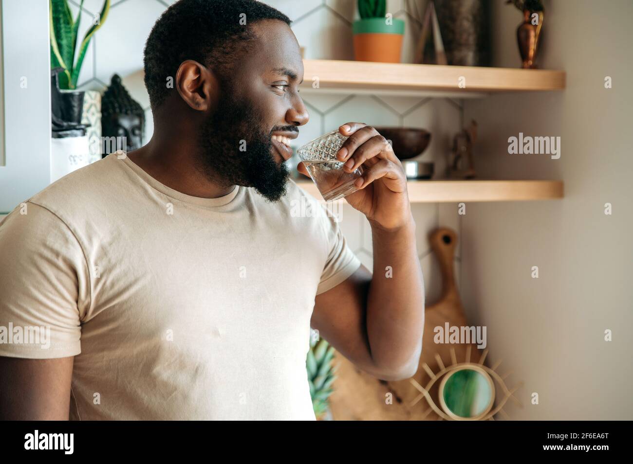 Afroamericano hombre atractivo beber agua pura, seguir un estilo de vida saludable. Guapo se siente sediento, beber la cantidad diaria de líquido puro, de pie en la cocina, concepto de salud Foto de stock