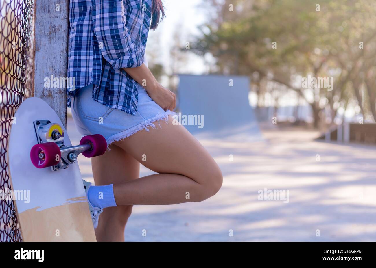 Las mujeres jóvenes que llevan pantalones cortos jeans y camisa de manga  larga apoyada en un polo de acero con un skate de surf al lado. Niña de pie  y relajarse después