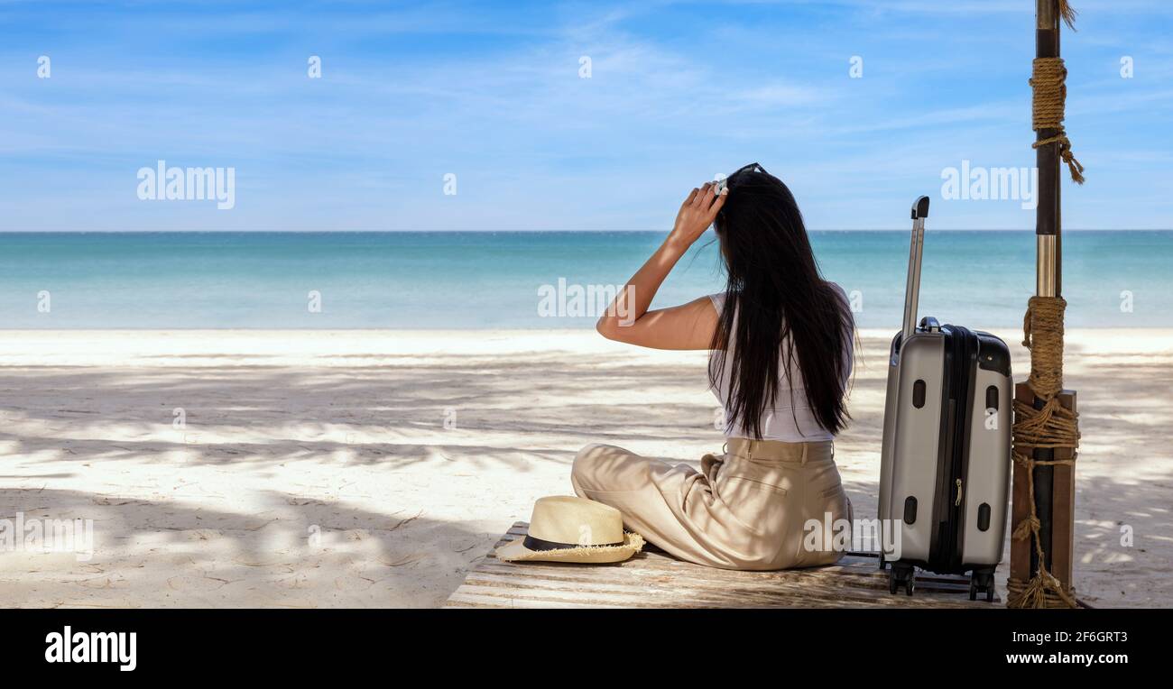 Joven mujer feliz con maleta de viaje sentada en la playa en verano. Mujer  de viaje mira al mar. Mujeres sentadas y relajadas con playa y backgrou del  cielo Fotografía de stock -