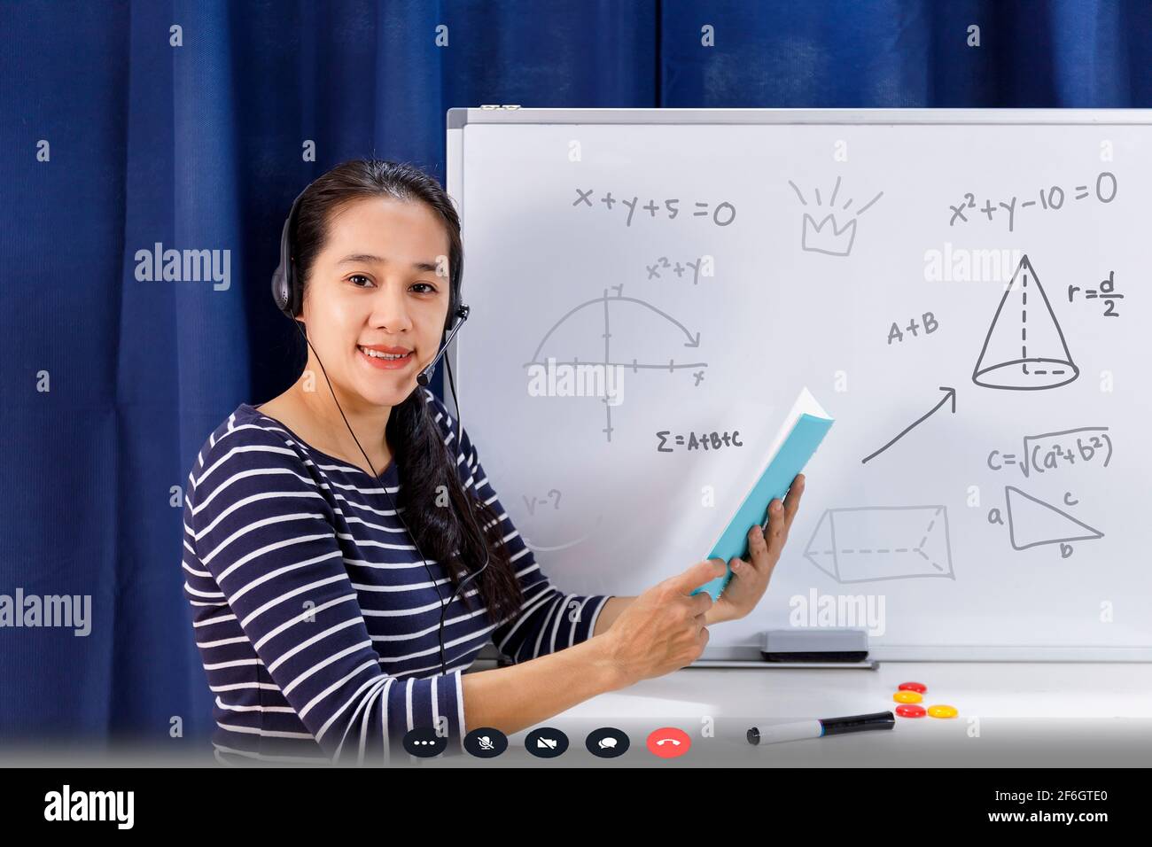 Una maestra de escuela asiática que trabaja desde casa enseña matemáticas  en línea a un estudiante que estudia desde casa. El profesor escribe en la  pizarra y habla en el auricular inter