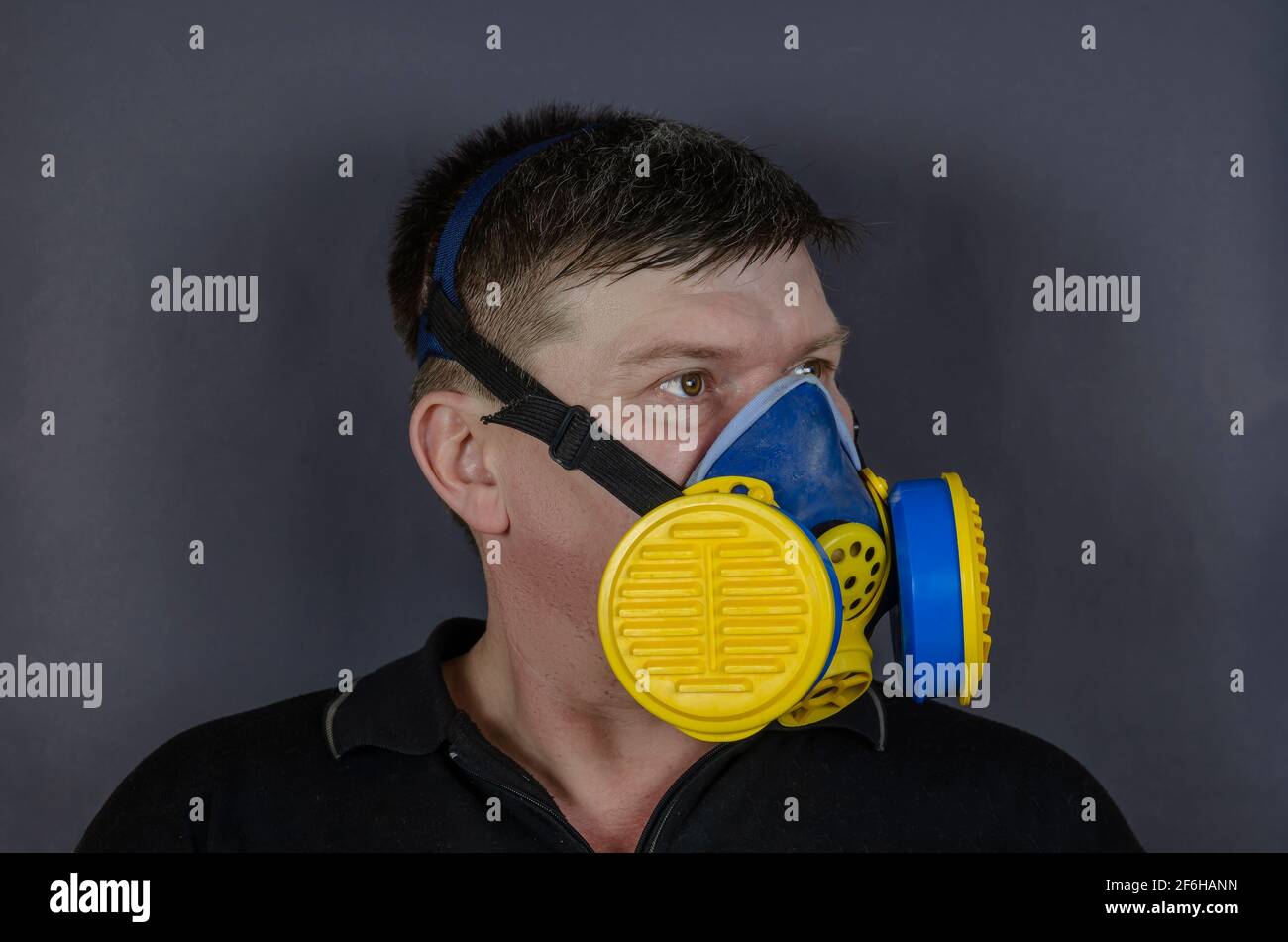 Retrato de un hombre en un respirador industrial sobre un fondo gris. Un hombre adulto con pelo corto usando un respirador azul y amarillo. Media máscara con Foto de stock