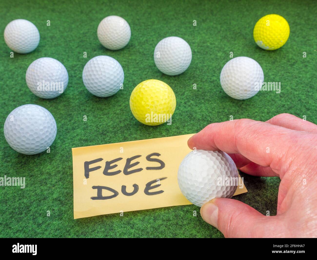 Mano de un hombre sosteniendo una pelota de golf en una nota de pago, entre otras bolas de golf en una superficie verde. Concepto de cargos por el uso de un club de golf / campo de golf. Foto de stock