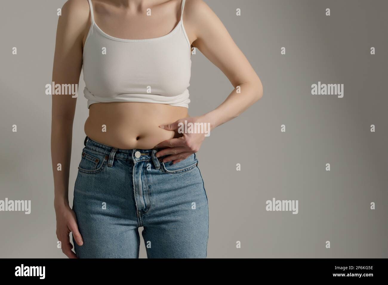 Mujer en jeans y camisa blanca pellizcando su vientre gordo. Figura de la mujer estudio de primer plano con fondo gris. Dieta y concepto de pérdida de grasa. Foto de stock