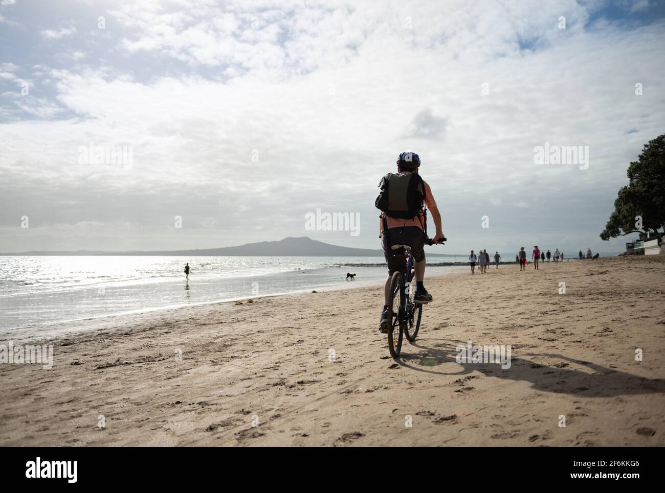 Un hombre en bicicleta en la playa de Milford con la isla Rangitoto en la distancia. Gente y perros desenfocados caminando por la playa. Foto de stock