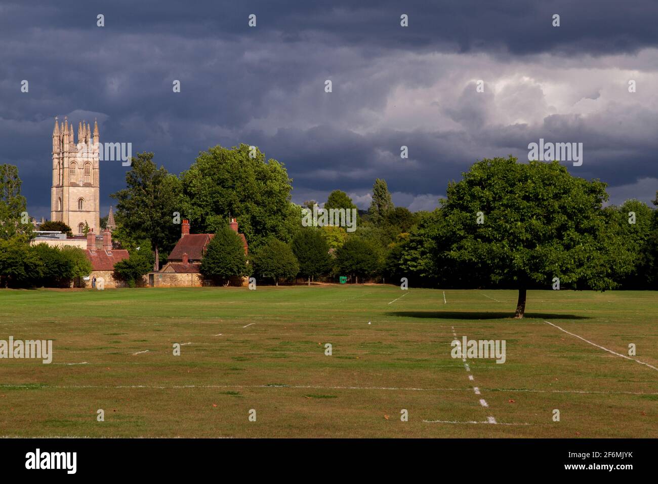 Tiro ancho de un campo deportivo rodeado de árboles y algunos pequeñas casas y la torre de la iglesia en el fondo en un día soleado con un cielo nublado Foto de stock