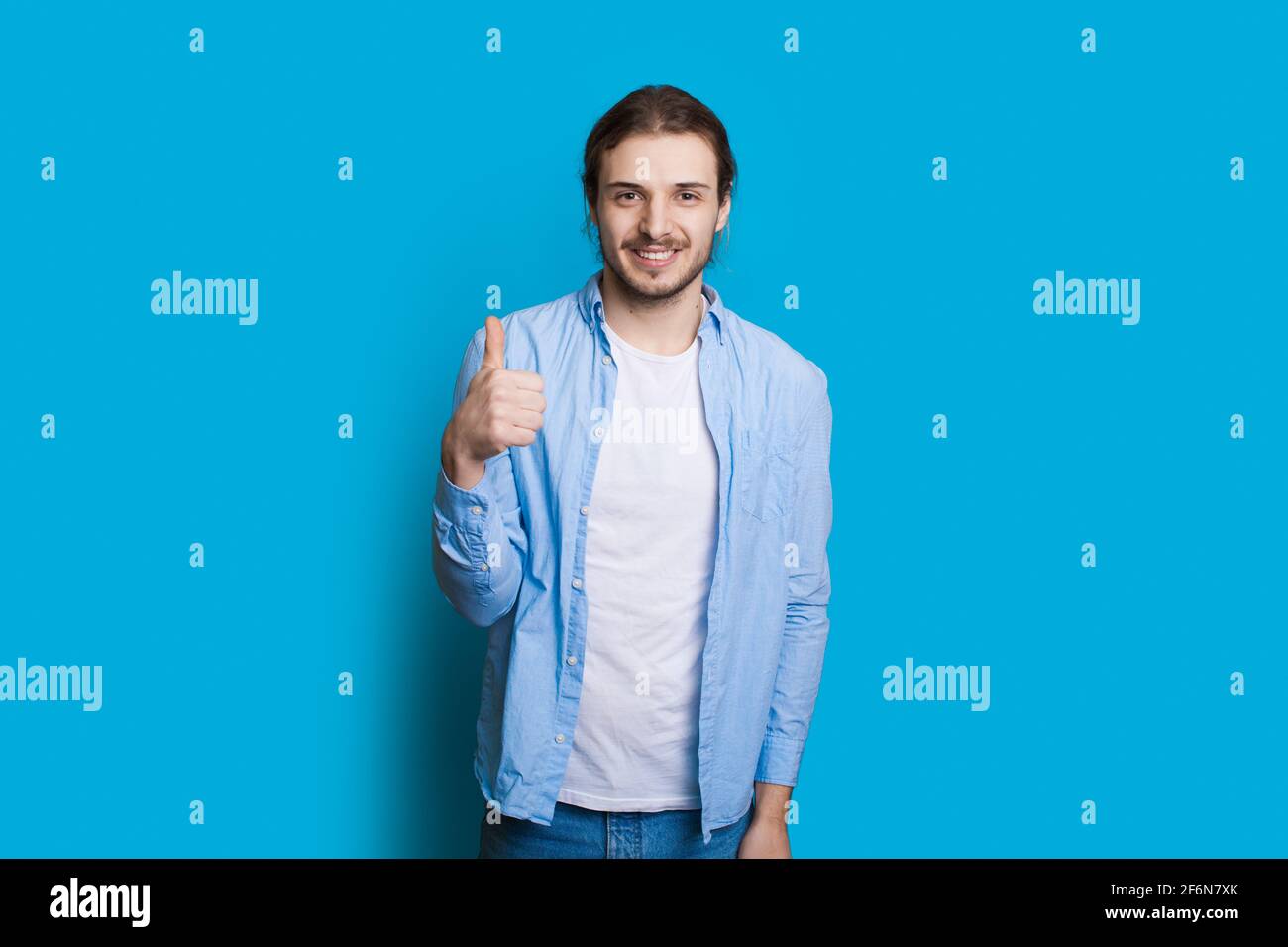 El hombre caucásico con barba y pelo largo está gestando el como signo en una pared azul del estudio Foto de stock