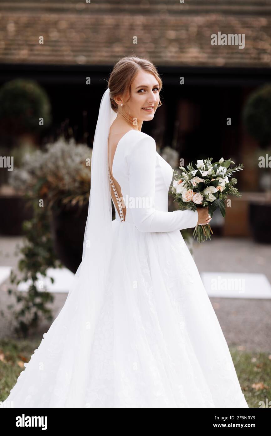 novia en un vestido largo blanco está de pie en el parque y la celebración de un ramo de de rosas flores y verdes. joven con peinado, maquillaje y Fotografía