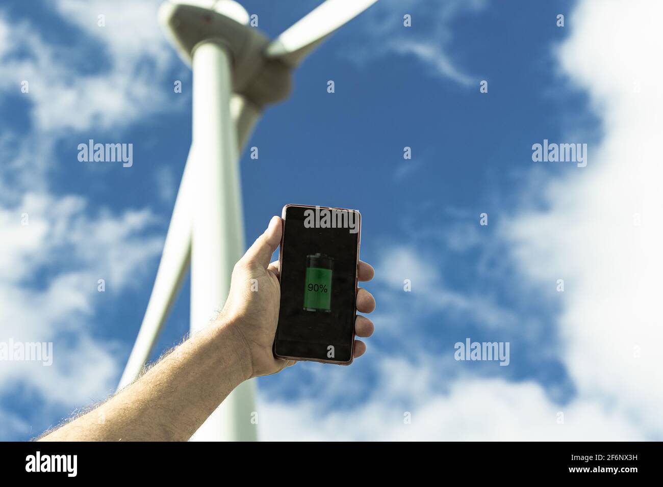 Retrato de mano de un hombre caucásico sosteniendo un smartphone, en el fondo un aerogenerador. Energía renovable, futuro y concepto de electricidad. Foto de stock