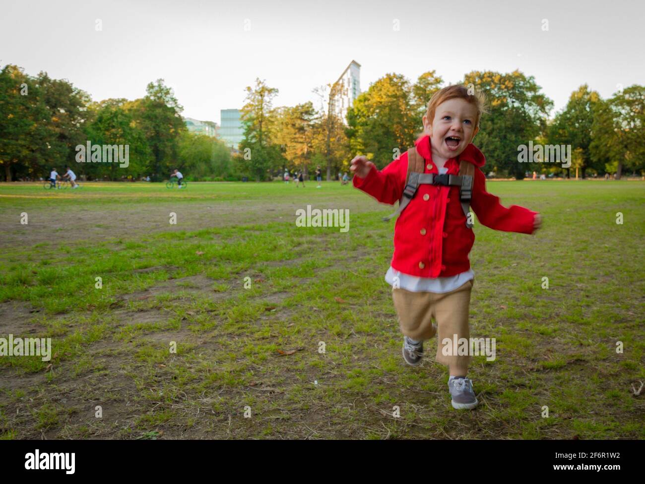 Un lindo, pelirrojo, niño con una chaqueta roja corriendo en un césped en un parque en una noche soleada Foto de stock