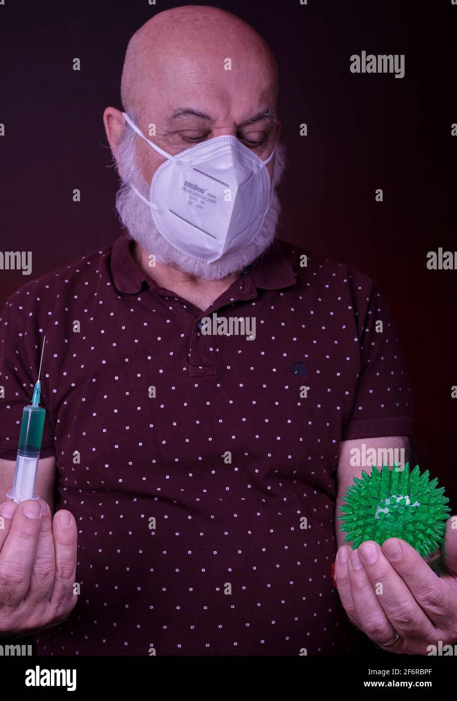 un hombre mayor con máscara protectora sostiene una jeringa de vacuna una mano y una bola verde que representa el virus de la corona en otro Foto de stock