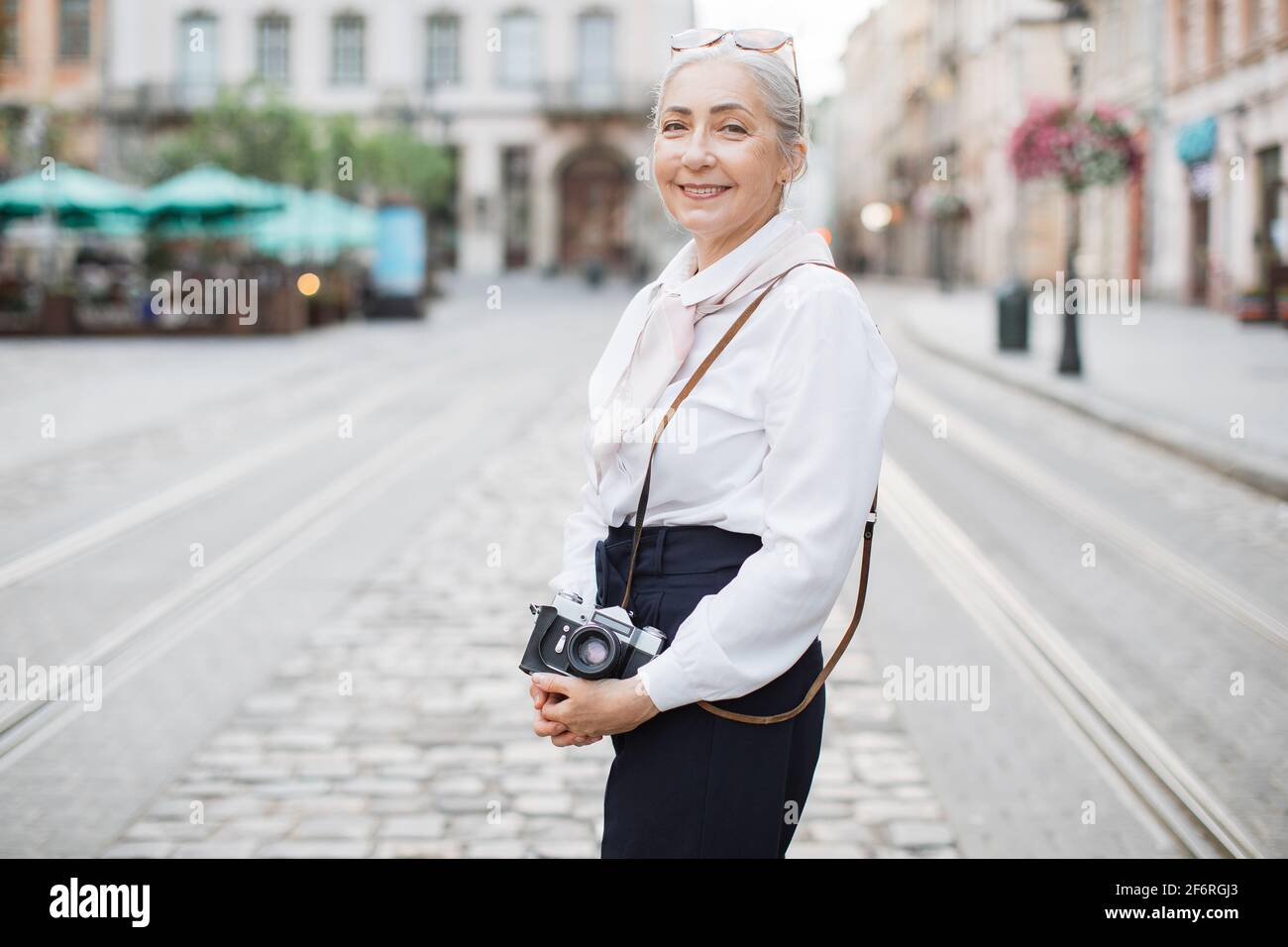 Mujer sonriente con ropa elegante que sostiene una cámara fotográfica retro está de pie al aire libre. Calle de la ciudad sobre fondo borroso. Mujer activa en la jubilación Fotografía