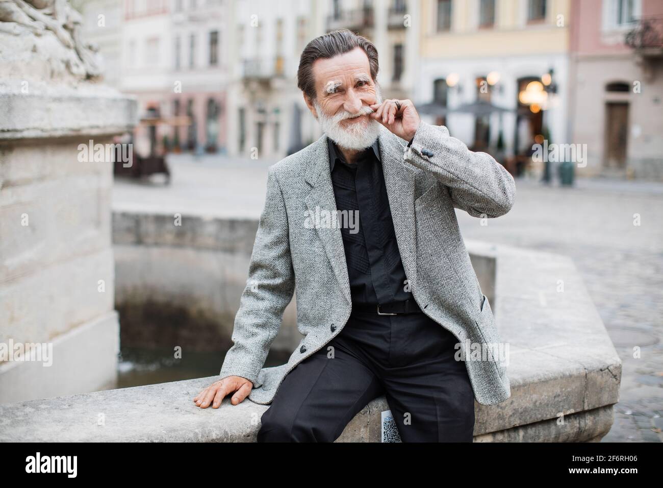 Hombre mayor barbudo vestido con ropa elegante posando cerca de la vieja  fuente de la calle de la ciudad. Pensionista sonriente relajándose al aire  libre y ajustando su bigote Fotografía de stock -
