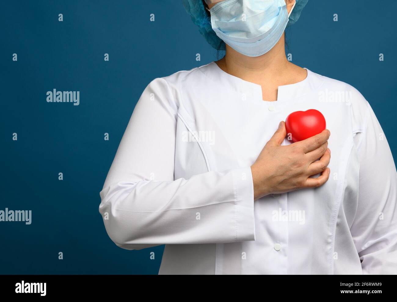 médico femenino en un abrigo blanco, una máscara se levanta y sostiene un corazón rojo sobre un fondo azul, el concepto de donación y amabilidad. Foto de stock