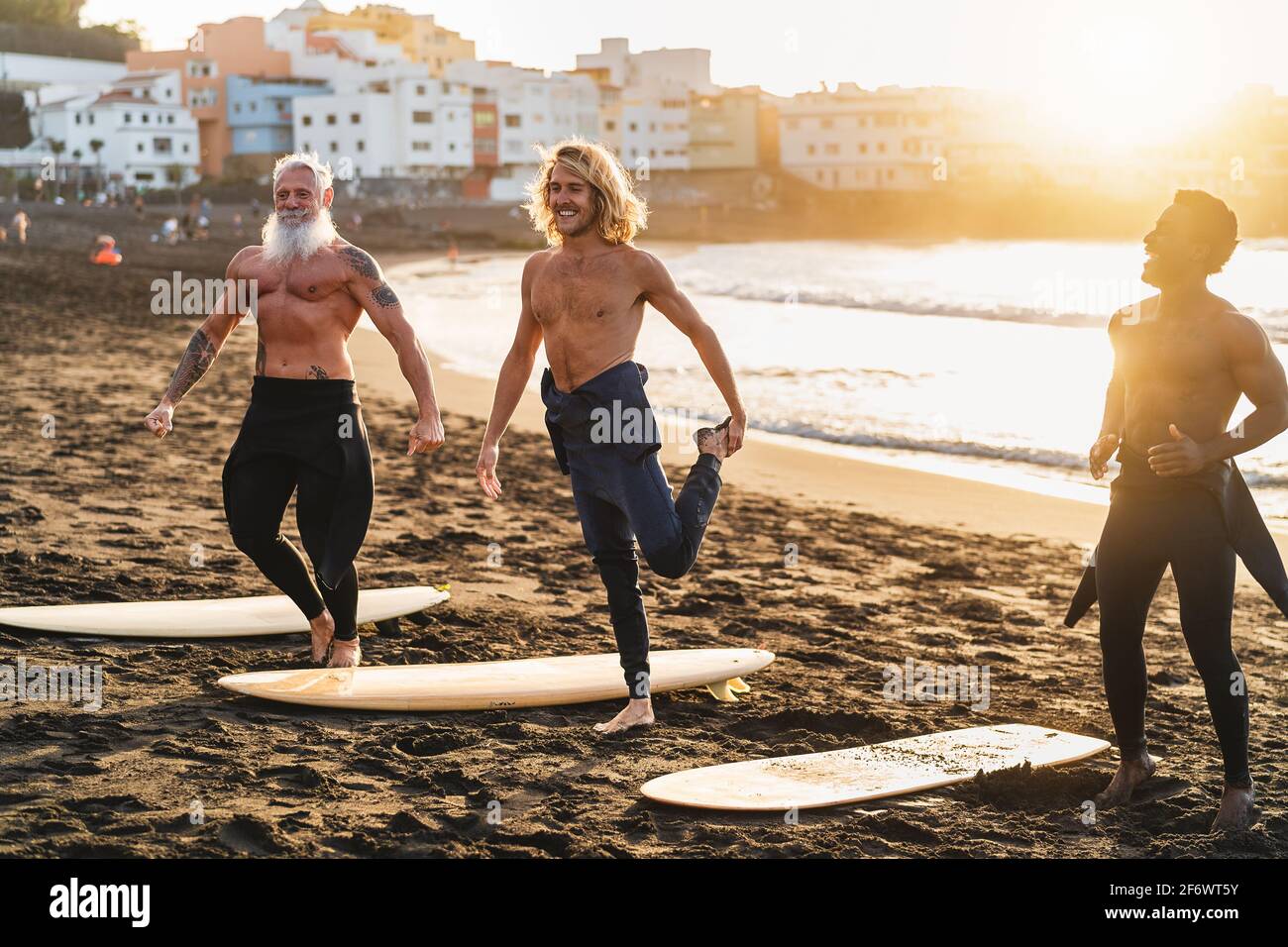 Felices surfistas con diferentes edades y carreras haciendo calor ejercicios antes de surfear durante la puesta de sol Foto de stock