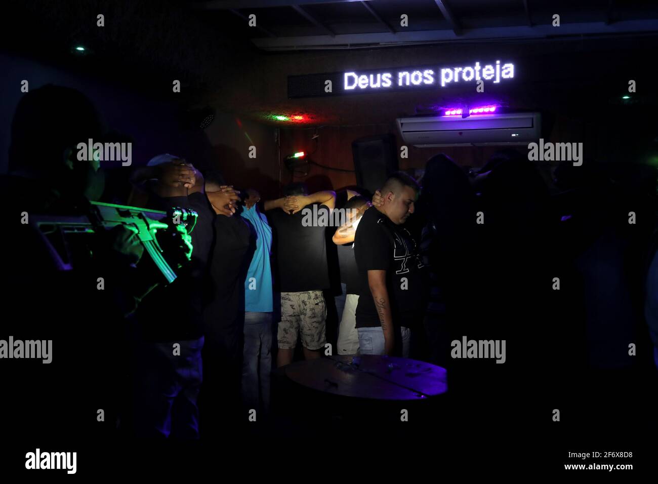 Las personas sin máscaras protectoras pusieron sus manos detrás de sus cabezas cerca de una luz de neón que dice "Dios nos protege" como un oficial de policía con un arma camina cerca en un club nocturno durante una operación de policía civil que aborda las violaciones de las restricciones COVID-19, En medio de un aumento de casos de enfermedad coronavirus y muertes en Sao Paulo, Brasil 2 de abril de 2021. Foto tomada el 2 de abril de 2021. REUTERS/Amanda Perobelli Foto de stock