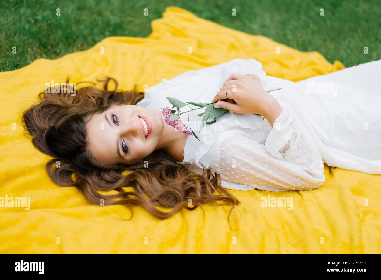 Arriba ver a la joven en un picnic, tumbada en una manta. Ella es feliz y sonríe una deslumbrante sonrisa blanca como la nieve y sostiene una flor en sus manos Foto de stock