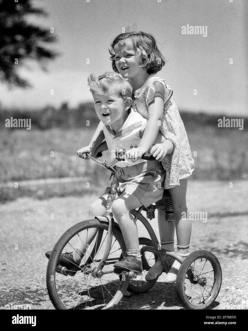 1930S NIÑO Y NIÑA HERMANO HERMANA MONTAR TANDEM EN TRICICLO CHICA  DIRIGIENDO EN LA PARTE TRASERA DE TRIKE NIÑO PEDRANDO RIENDO TENER  DIVERSIÓN - J6278 HAR001 HARS RURAL HOGAR COPIA DE VIDA