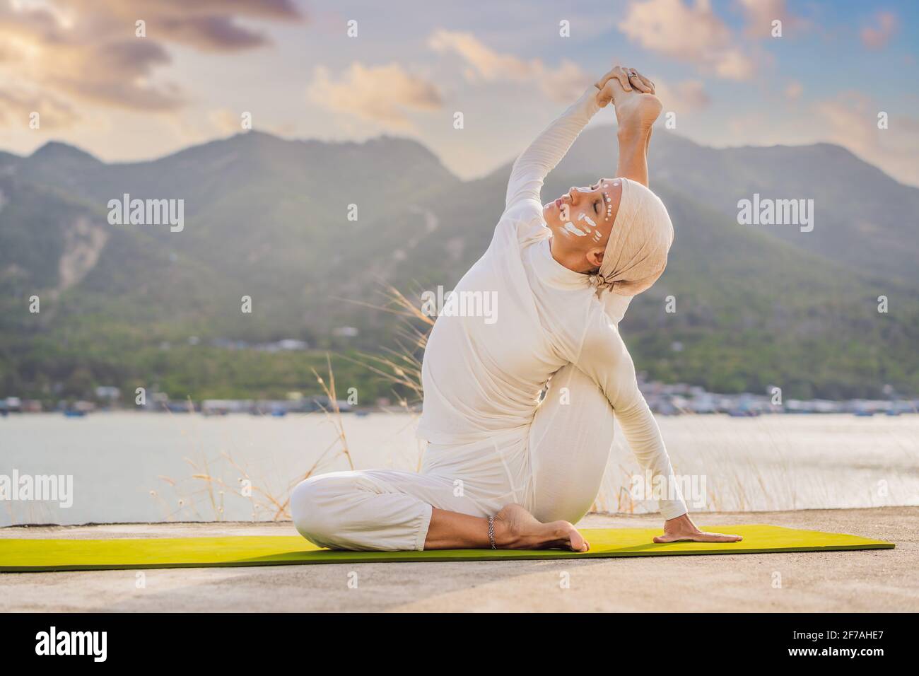 Kundalini yoga mujer con ropa blanca y turbante practica yoga kundalini en fondo del mar, las montañas y la puesta de sol. Lucha contra la pintura de cara Fotografía de stock -
