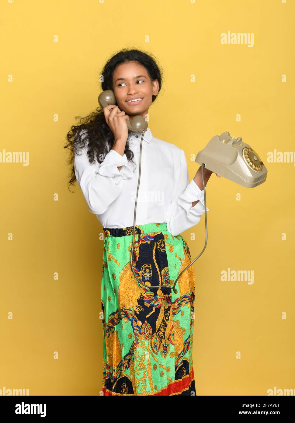 Joven dominicano de moda en falda de colores brillantes y elegante blusa blanca de pie hablando en un teléfono de época sobre un fondo de estudio amarillo Foto de stock