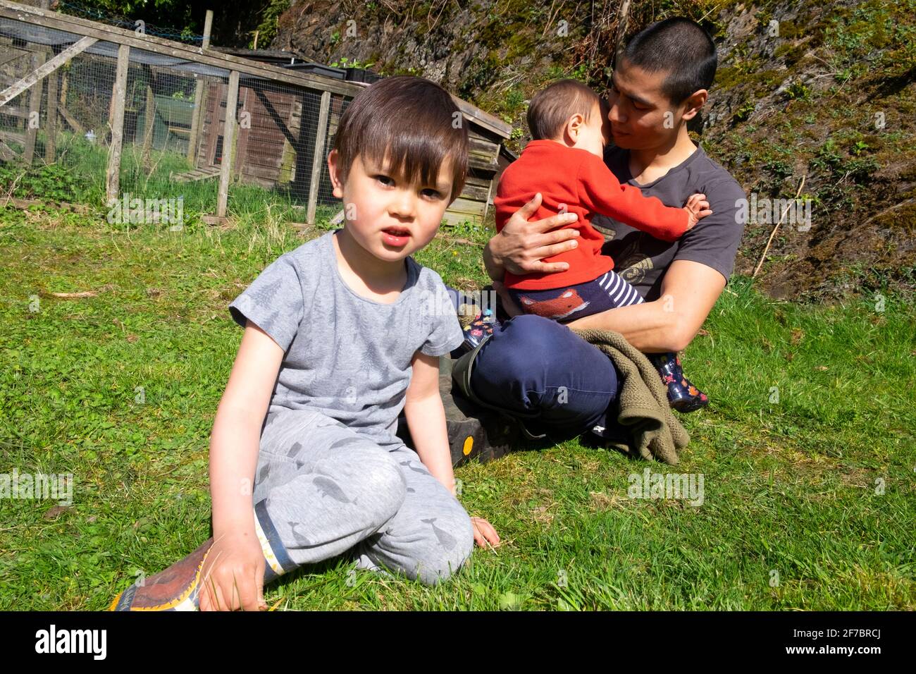 Raza mixta chico asiático mirando y hombre padre padre sentado Afuera en la hierba en primavera sosteniendo al niño Carmarthenshire Wales REINO UNIDO KATHY DEWITT Foto de stock