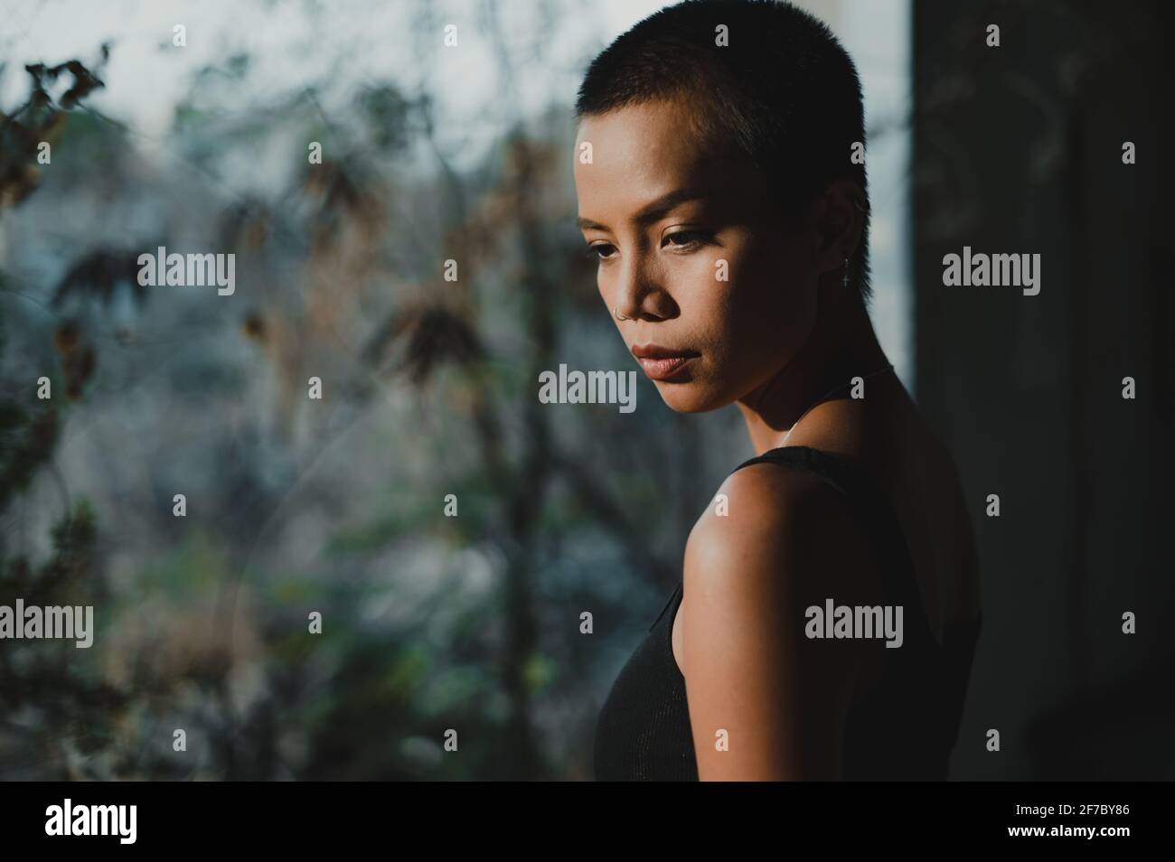 Retrato de una joven mujer asiática de pelo corto de pie junto a una gran ventana mirando hacia abajo, con árboles fuera de foco en el fondo. Foto de stock