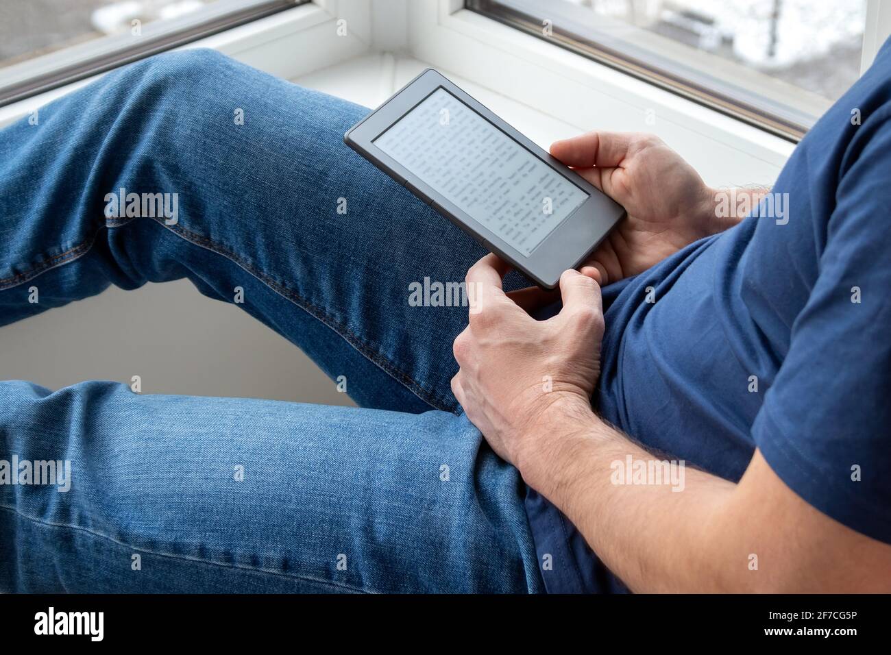 Un hombre en una camiseta azul y jeans sostiene un libro electrónico e-Reader en sus manos. Leyendo libros en casa junto a la ventana. Enfoque selectivo Foto de stock