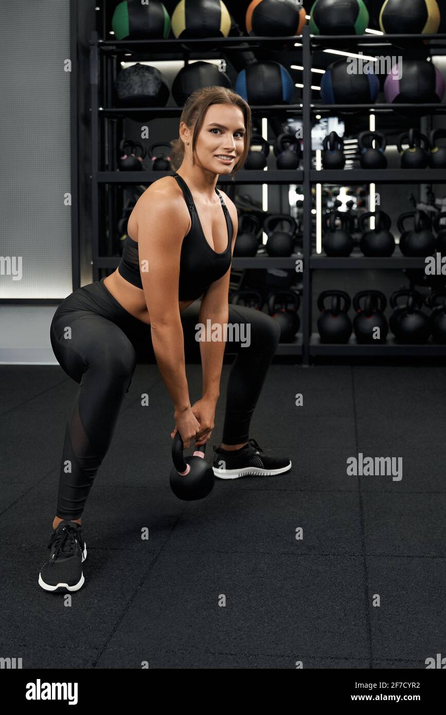 Vista lateral de la joven mujer sonriente en la hermosa ropa deportiva que  sostiene las campanas de las hervidores y hacer ejercicio para todo el  cuerpo. Concepto de entrenamiento y bombeo de